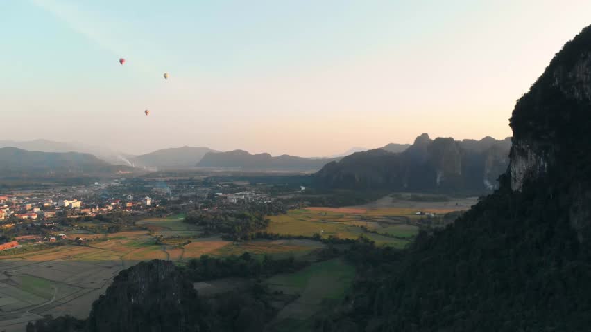 Aerial: Vang Vieng backpacker travel destination in Laos, Asia. Hot air balloons at sunset over scenic cliffs and rock pinnacles, rice paddies valley, stunning landscape.
