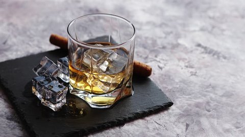 Glass of whiskey with ice cubes and cigar placed on top of stone serving plate. Stone marble background.