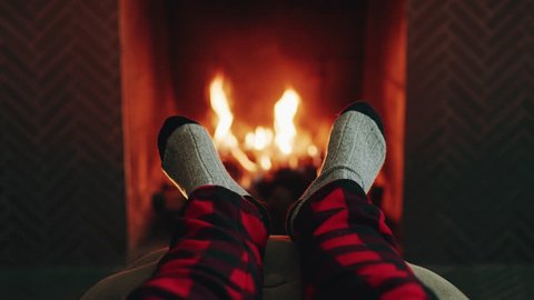 Relaxation. Someone puts their feet up on a foot rest by the fire. They’re wearing cozy socks, and plaid pants. It’s incredibly cozy.  วิดีโอสต็อก