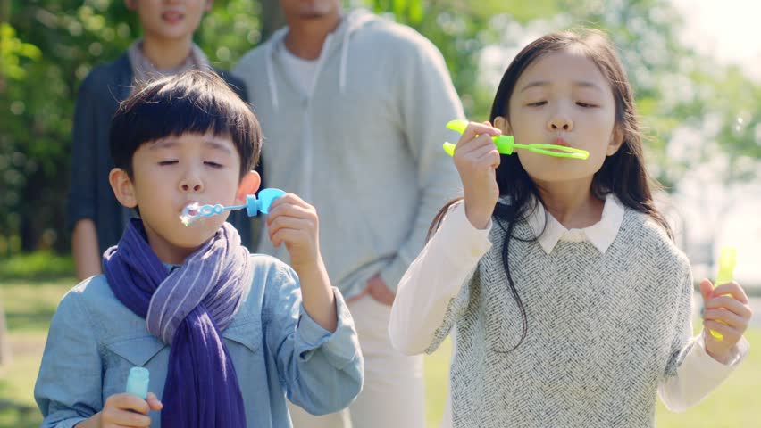 Two little asian children boy and girl playing outdoors blowing soap bubbles with parents watching from behind. | Shutterstock HD Video #1022674519