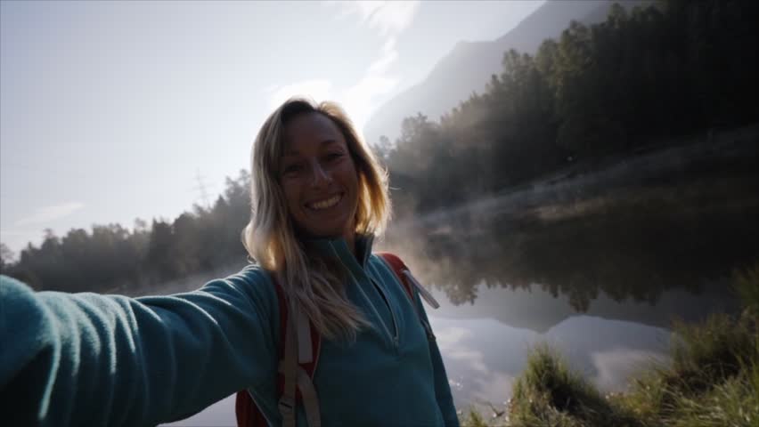 Cheerful young woman taking a selfie portrait by a beautiful mountain lake enjoying day hike 