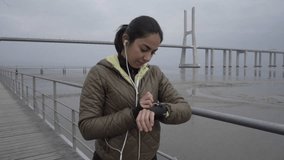 Smiling sporty young girl setting sport watch on seaside. Attractive brunette woman preparing to running. Concept of sport