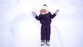 Point of view video of cute little funny baby girl laying in snow happily and waving her hand into camera smiling. Christmas fun and happy winter games concept. Slow motion top view footage