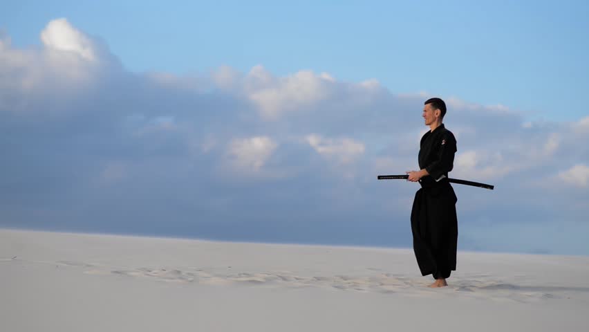 Concentrated man, in traditional Japanese clothes, with sword, katana, is training martial arts in desert during sunset - samurai on the blue cloudy sky background. 