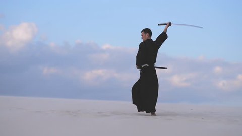 Concentrated man, in traditional Japanese clothes, with sword, katana, is training martial arts in desert during sunset - samurai on the blue cloudy sky background. 