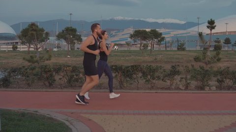 Two joggers are performing his morning jog in park area in city near mountains. Man and woman are running together and enjoying picturesque landscapes, side view