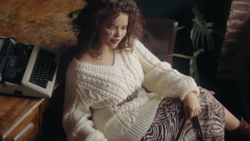 Young beautiful fashionable woman wearing cozy white knitted sweater, animal print skirt, belt, holding small brown bag, posing at loft interior
