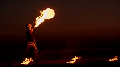 Cool fire show artist standing between bowls with burning liquid breathes fire in dark air, performing amazing stunts - slow motion