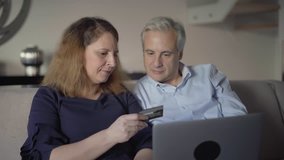 Positive middle aged couple paying online. Mid adult man and woman using computer at home, inserting credit card data for transaction. Online payment concept