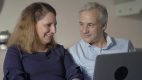 Positive mid adult couple with laptop discussing new online app or service. Middle aged man and woman sitting on couch, talking to each other and pointing at monitor. Online app concept
