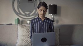 Serious freelancer with laptop focused on work. Beautiful young mix raced woman working on computer at home, sitting on couch and typing. Working at home concept