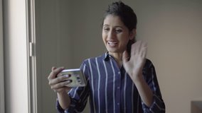 Positive Latin girl saying Hello to smartphone screen. Young woman in blue casual shirt holding phone, waving hand, greeting and talking to screen. Video call concept