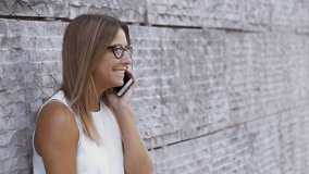 Beautiful happy girl talking by cell phone. Attractive young woman leaning at brick wall and using smartphone during phone call outdoor. Communication concept