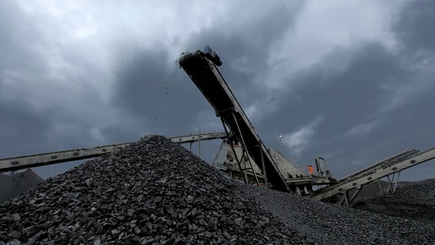 An overview of coal falling from the moving belt conveyor into the huge pile at the coal mine with coal miners working on the background