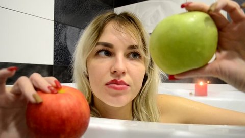 Beautiful girl bathing in the bathroom and starts watching a big green and red apple. 
The girl can not choose which apple she wants to eat