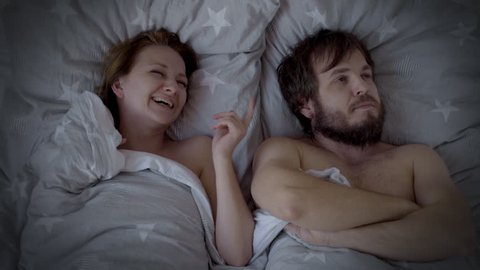 Top view of Upset bearded Man in bed having problems with Sex, impotence. Couple lying in Bed - Husband suffering problems with masculine Health, Wife laughs.