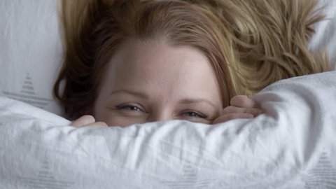 Closeup of Lovely young Woman Lies in Bed Covered with Blanket. Top view of Girl is Shy and Cover of her Face with Blanket. Female Smiling and Looking at Camera.