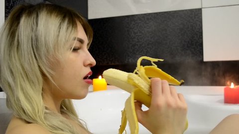 An attractive woman sits in the bathroom and brushes the big yellow banana and starts with the pleasure of eating it. Romantic atmosphere in the bathroom