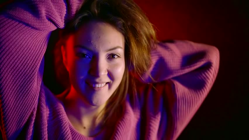portrait of a pretty cheerful young girl in a dark room dancing in the light of neon blue and red. dressed in a pink knitted sweater