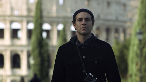 Outside of the Coliseum in Rome, an Italian man with a camera around his neck walking in a park. Wide shot on 8k helium RED camera.