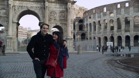 Happy, cheerful Italian couple walk together while looking at the buildings, including the Coliseum in the background, with soft natural light. Wide shot on 4k RED camera.