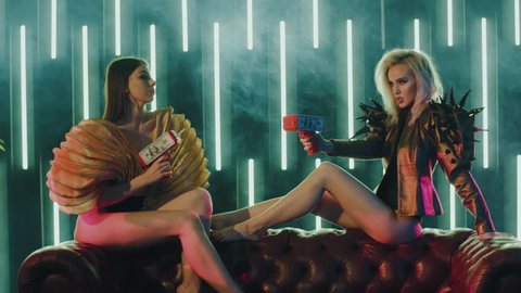 Two sexy women sitting on couch in stylish dresses and shooting money from money guns. Dollar bills flying in a room with green neon light. Shoot with RED RAVEN camera.