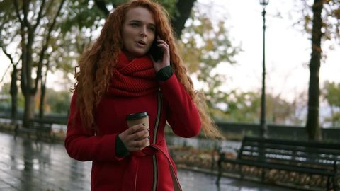 Young serious girl talking on her mobile phone on while walking in the autumn park. Curly haired woman in red coat using her smartphone and holding paper cup with coffee. Woman walking and talking