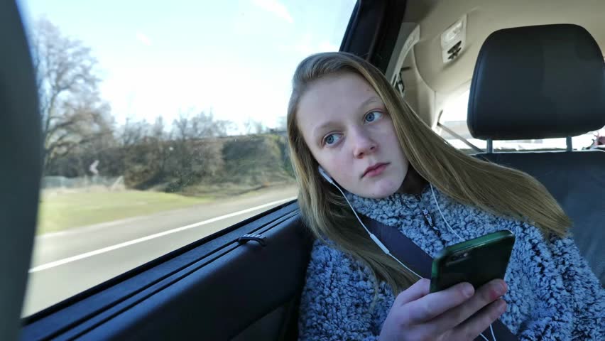 A cute blonde teenager riding in a moving car, leans her head against the window and types on her smart phone while wearing earphones