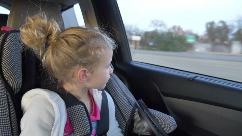 A cute little blonde girl sits in a car seat and looks out the window of a moving vehicle