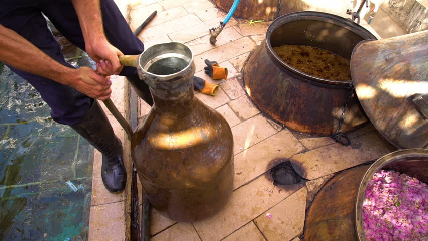 Hand held, medium close up shot of man pouring kettle of water into a drum kettle filled with rose petals.