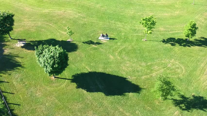 Aerial flight above two young people seating on the bench in the sunny summer green grass spacey area. Couple.