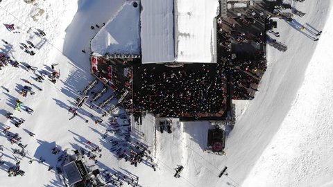 Skiers and snowboarders enjoying apres ski party outside a bar and chairlift with the backdrop of snowy Alps slopes