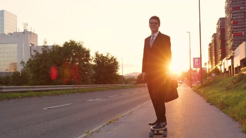 SLOW MOTION, LENS FLARE: Smiling Caucasian businessman skateboarding down the empty sidewalk at sunset. Cheerful yuppie riding his electric longboard cruising home on a picturesque summer evening.
