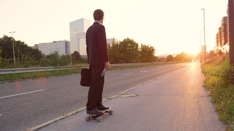 SLOW MOTION, SUN FLARE: Young man in a black suit riding his skateboard down the empty pavement and into the golden sunset. Unrecognizable sporty businessman going home on his cool electric longboard.