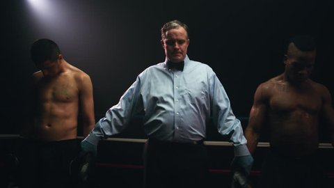 The referee stands with two boxers after a match and announces the boxing champion. He raises his arms in excitement and smiles at the camera