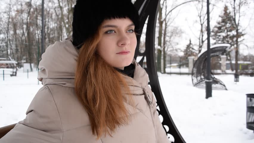 Young red-haired girl sitting on a wooden bench in the winter Park
