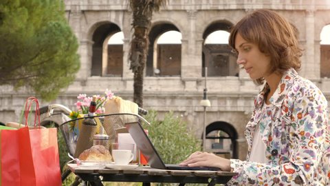 Young woman writing and working on her laptop computer sitting at the table outside in a bar in front of the Colosseum in Rome. Elegant beautiful dress and colorful shopping bags.