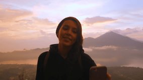 Young Smiling Mixed Race Tourist Blogger Girl Recording Selfie Video from the Top of the Mountain Using Smartphone with Beautiful Sunrise on Background. 4K Slowmotion Travel and Freedom Lifestyle