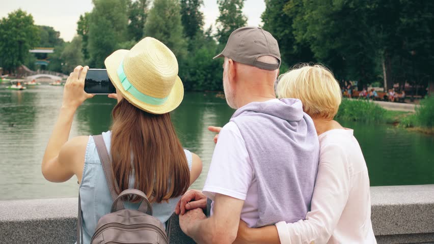 Back view of happy family enjoying summer vacation together in Moscow, Russia. Seniors with young teenage daughter having great time standing by lake in park laughing and chatting sightseeing