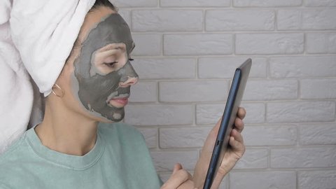 Woman using tablet while waiting drying mask. Head wrapped in towel.