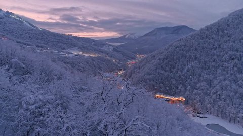 Aerial drone establishing shot of white winter Rosa Khutor resort in Sochi. Snow covered trees. Evening illumination. After sunset. Flying low above snow forest