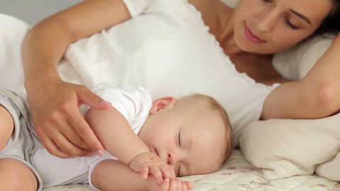 Happy mom is lying with her newborn sleeping baby in bed, she admires the baby, gently strokes and kisses him.