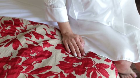 Close-up of the bride in a satin negligee sitting on the bed with red flowers in the hotel, hand close-up.