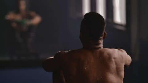 Over the shoulder shot of a sweaty African American athlete sitting and peering out a window in a boxing gym, his image reflected in the mirror