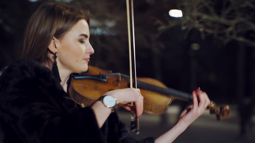A charming girl is playing the violin in a black coat in the evening in the street. Camera motion around