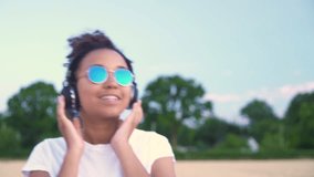 Slow motion follow shot video of beautiful mixed race African American girl teenager young woman in white T-shirt and blue sunglasses walking listening to music on wireless headphones 