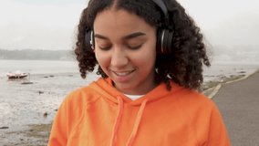 Follow shot video of beautiful mixed race African American girl teenager young woman wearing orange hoodie, walking by a harbor and listening to music on wireless headphones using her smart cell phone