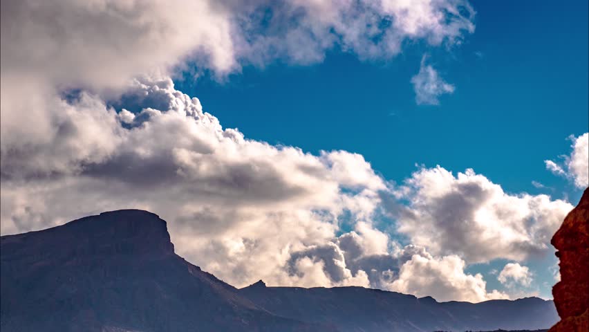 Bright blue sky with soft white clouds over a rocky and desolate landscape. Purple and red hue on the dry terrain from the sunlight. TILT DOWN to reveal a wide desert valley. Motion time lapse.