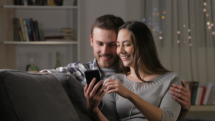 Happy couple sharing earphones watching videos online on a smart phone sitting on a couch at home in the night Royalty-Free Stock Footage #1022901790