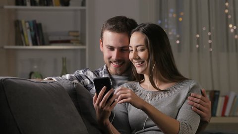Happy couple sharing earphones watching videos online on a smart phone sitting on a couch at home in the night